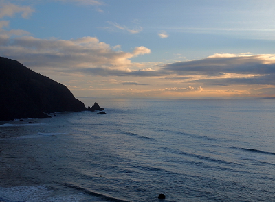 [With the dark mountain to the left and the ocean in the foreground, the yellow and white clouds streak across the background with blue skies still visible at the top of the image. Calm waves roll to the shore.]